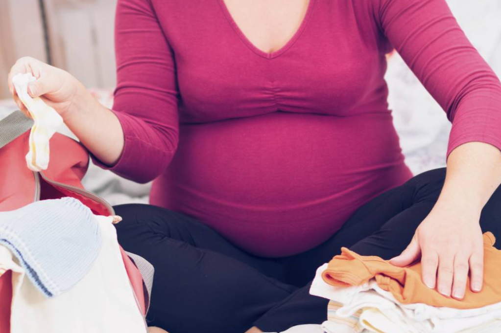 Беременная женщина в бордовом свитере складывает детские вещи